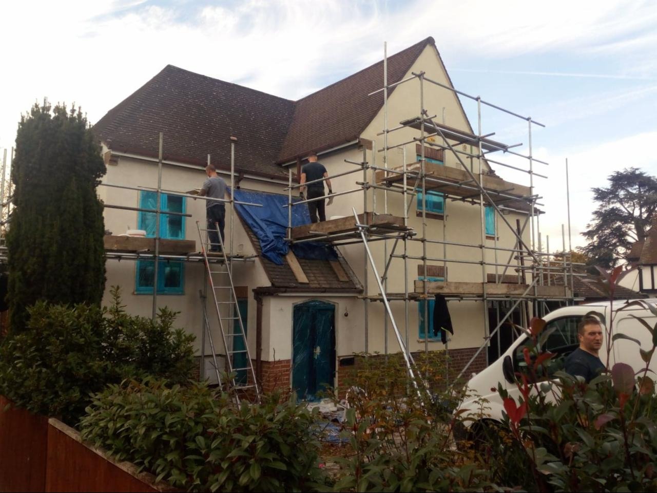 Dwóch pracowników na rusztowaniu i drabinie, przygotowujących elewację dwupiętrowego domu z brązowym dachem, okna zabezpieczone folią, widoczny biały samochód dostawczy.