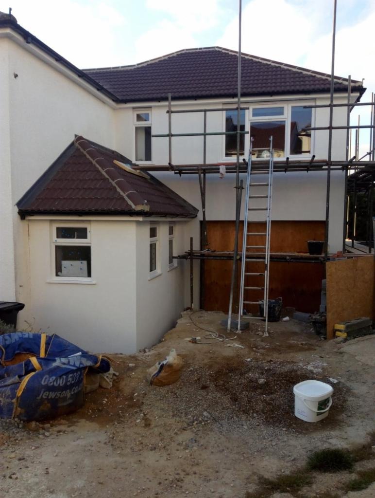 Dom w trakcie prac elewacyjnych: świeżo otynkowana ściana, rusztowanie, drabina, materiały budowlane na ziemi.