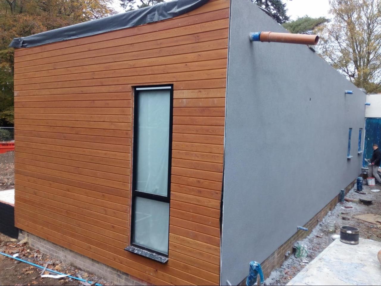 Budynek z elewacją częściowo wykończoną drewnianą okładziną i szarą strukturą, widoczne rury odprowadzające wodę i okno z czarną ramą, teren budowy.