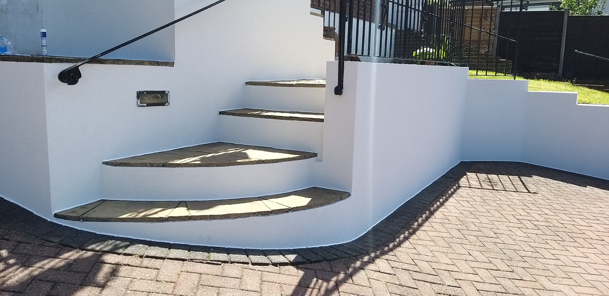 Białe, otynkowane schody zewnętrzne z kamiennymi stopniami i czarną balustradą, prowadzące do domu, z widoczną kostką brukową na podjeździe.