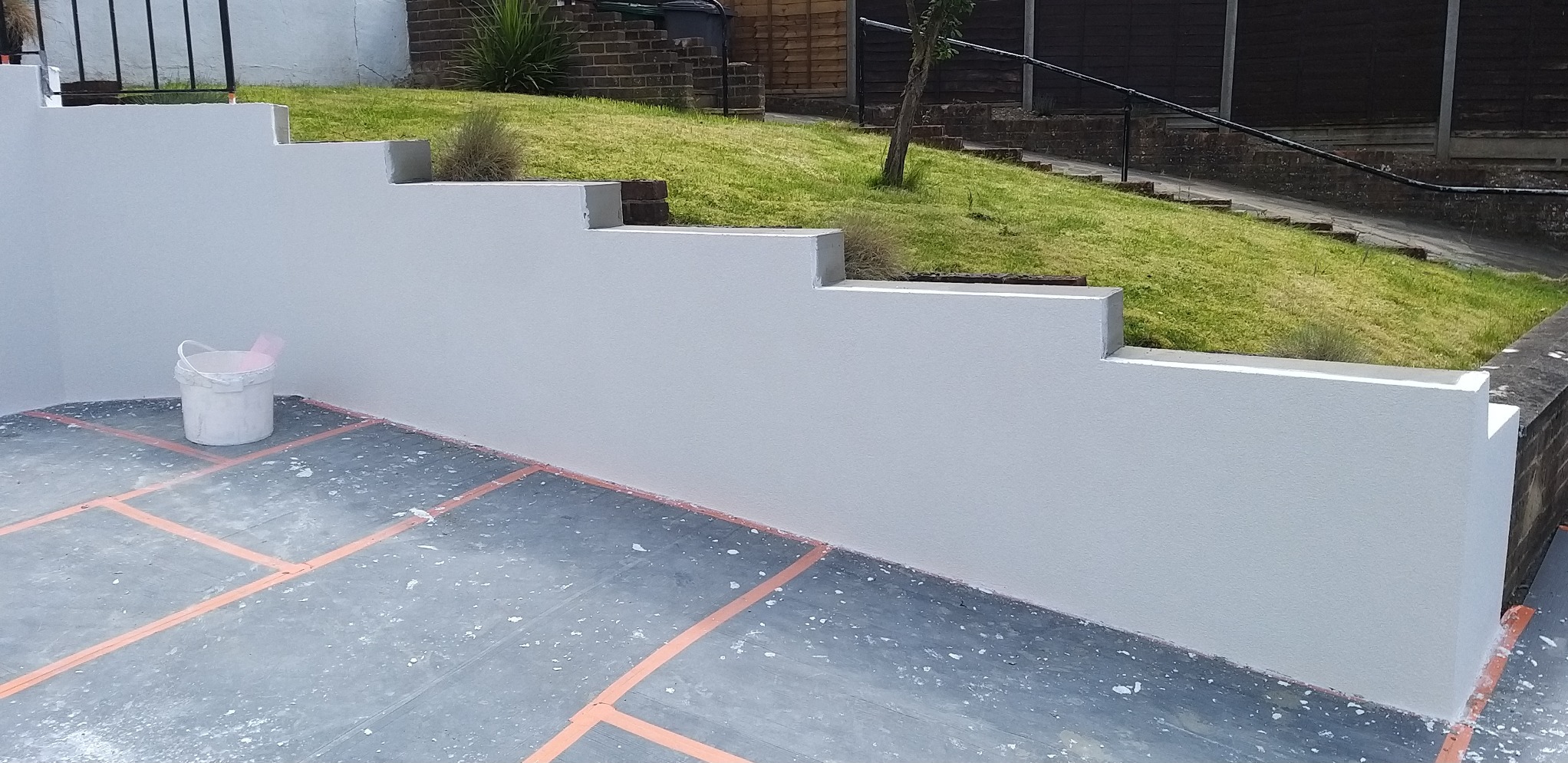 Świeżo pomalowany, schodkowy murek elewacyjny na zewnątrz domu, z wiaderkiem farby i zabezpieczonym podłożem.