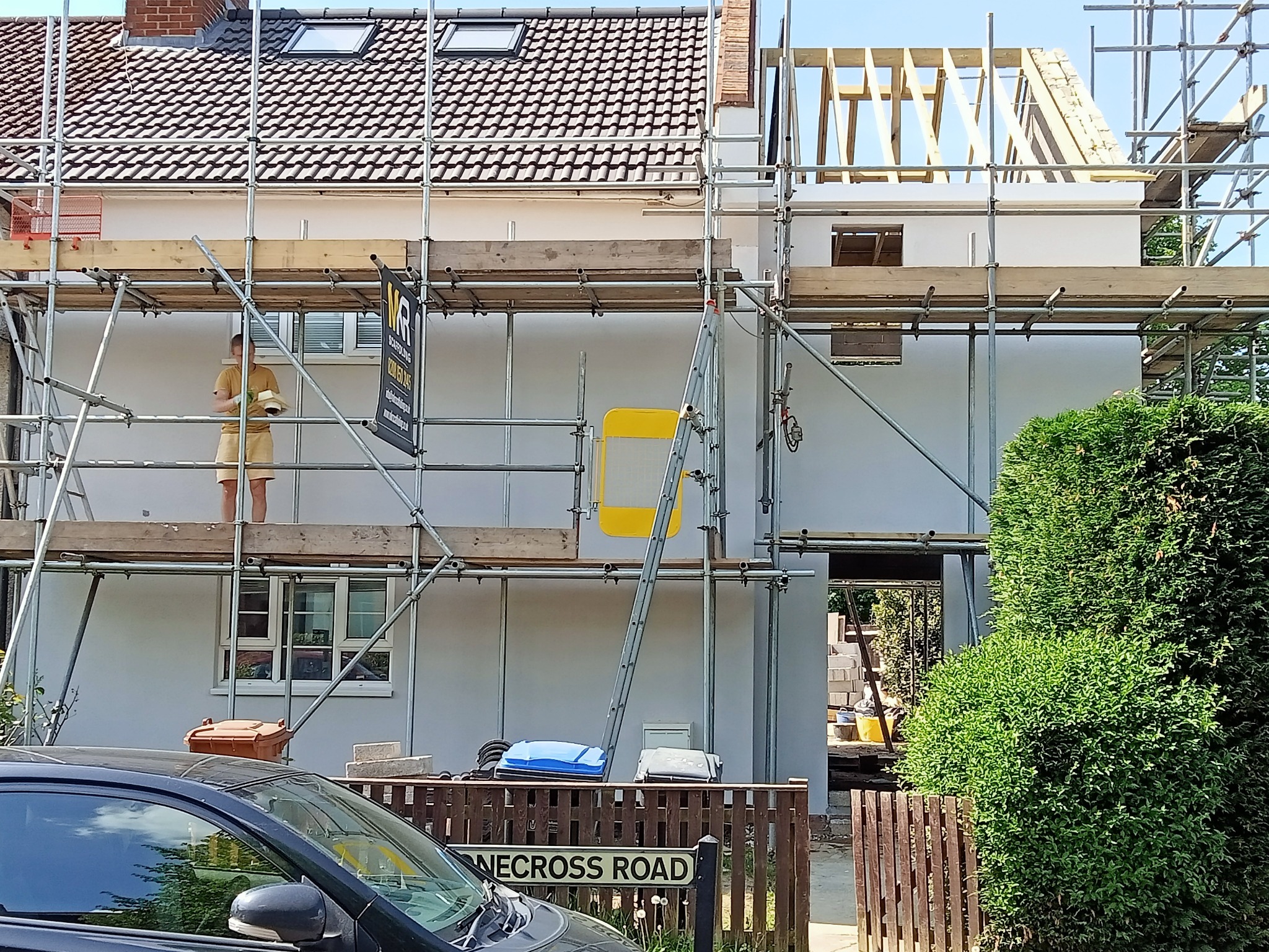 Dom jednorodzinny w trakcie prac elewacyjnych, widoczne rusztowania, częściowo wykonana konstrukcja dachu, pracownik na rusztowaniu i ulica z zaparkowanym samochodem.