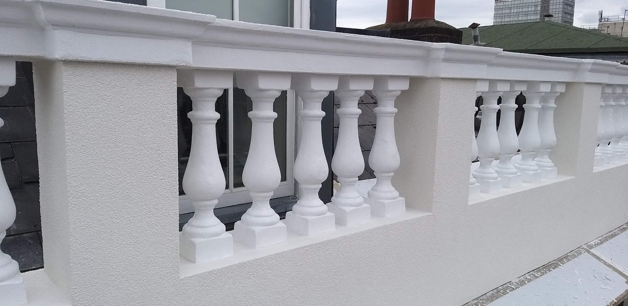 Biała balustrada z kolumnami na balkonie z tynkowaną elewacją o ziarnistej fakturze, widok na budynki w tle.
