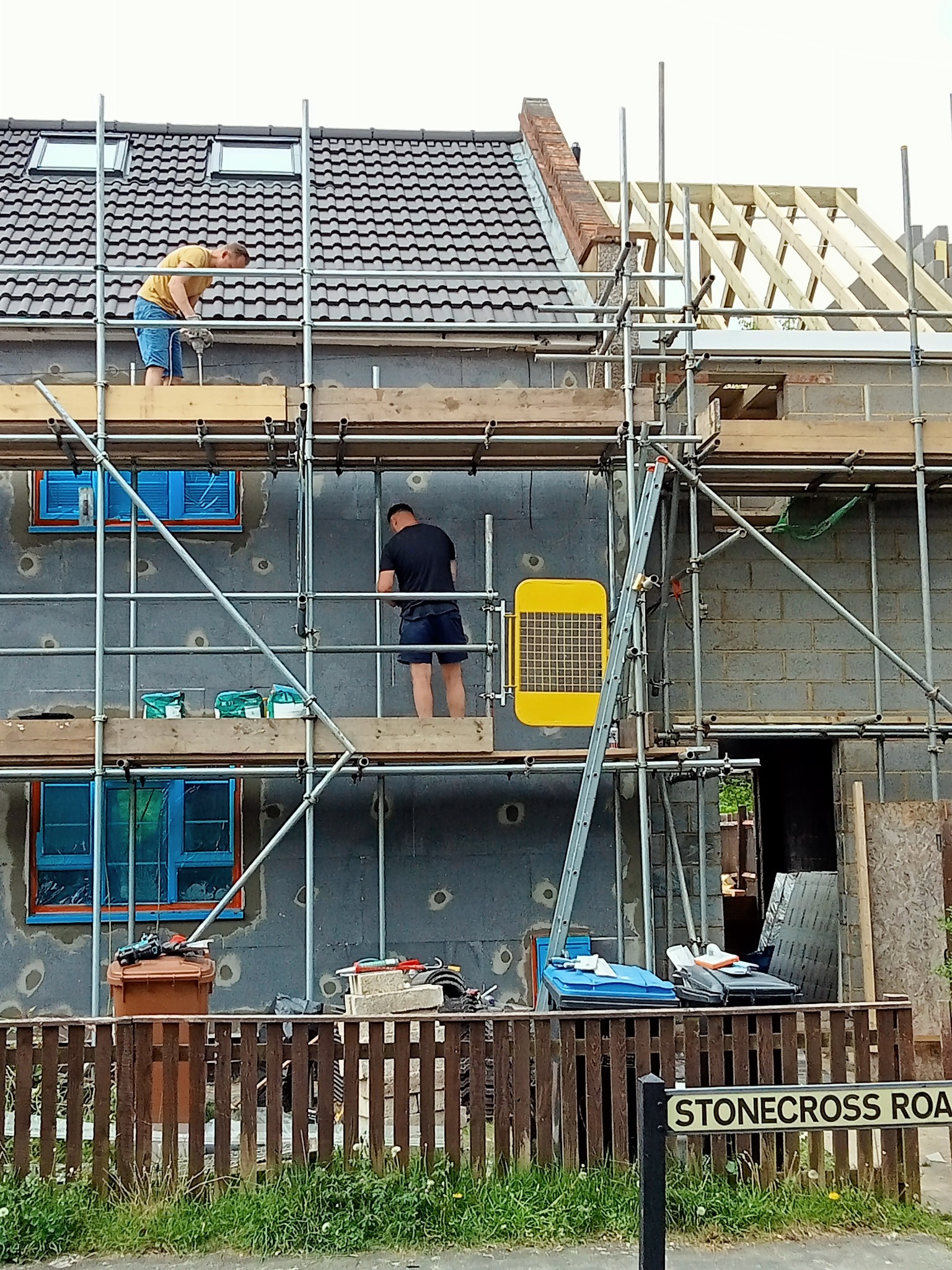 Prace na rusztowaniu przy ocieplaniu budynku z szarym styropianem, widoczne okna zabezpieczone folią i budowa dachu w tle.