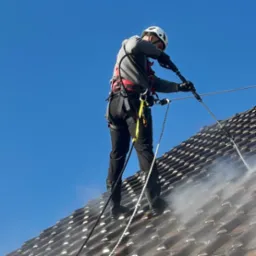 Pracownik w kasku i uprzęży czyści dach z brązowej dachówki za pomocą myjki ciśnieniowej. Widoczne liny asekuracyjne i strumień wody pod ciśnieniem.