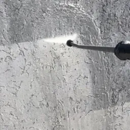 Czyszczenie szarej, teksturowanej elewacji strumieniem wody pod ciśnieniem z dyszy, widoczne ślady wilgoci po czyszczeniu.