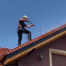 Odnova Masters Szymon Cichowlas - Pracownik w kasku i uprzęży myje dach z czerwonej dachówki za pomocą myjki ciśnieniowej na tle błękitnego nieba. Widoczny fragment elewacji budynku.
