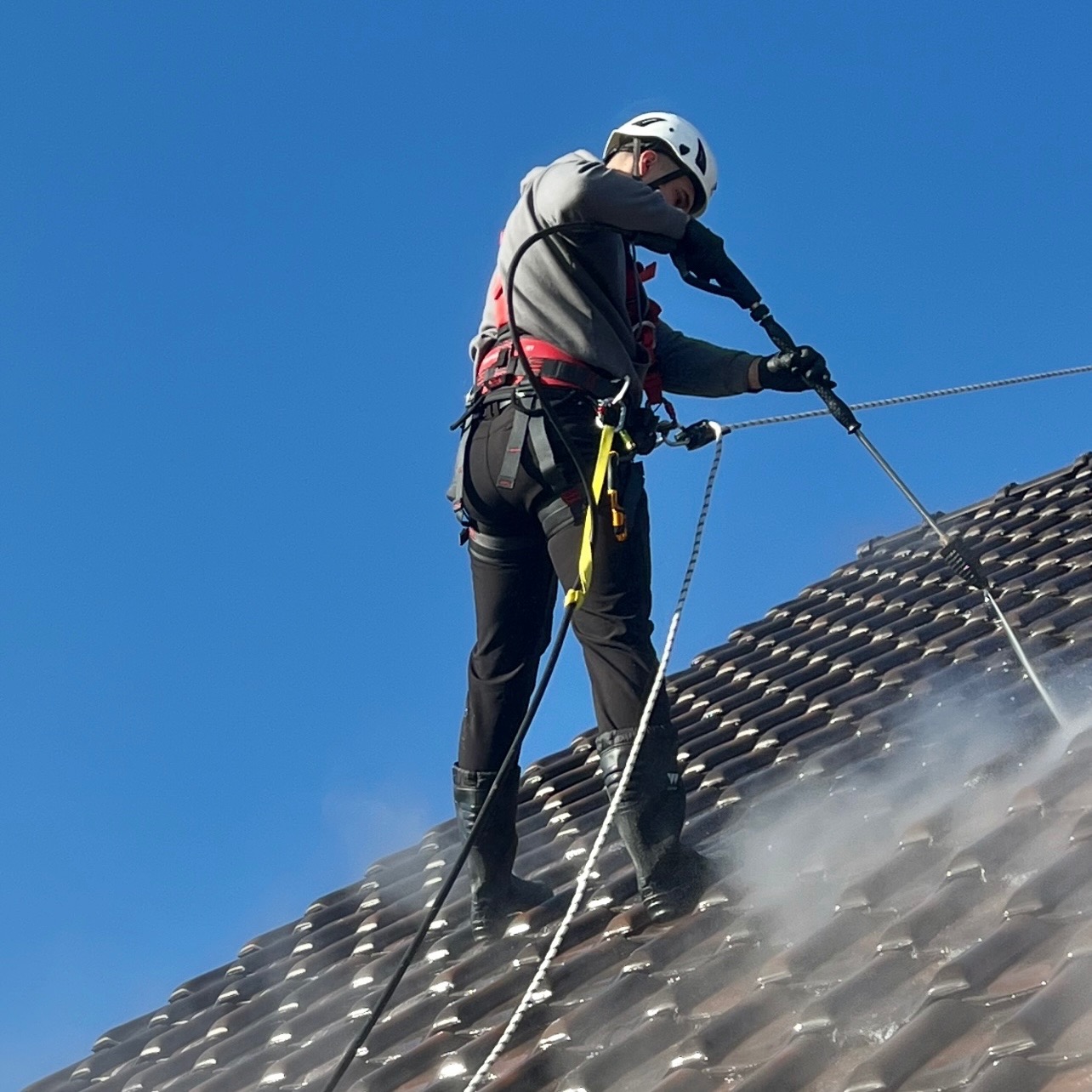 Pracownik w kasku i uprzęży czyści dach z brązowej dachówki za pomocą myjki ciśnieniowej. Widoczne liny asekuracyjne i strumień wody pod ciśnieniem.
