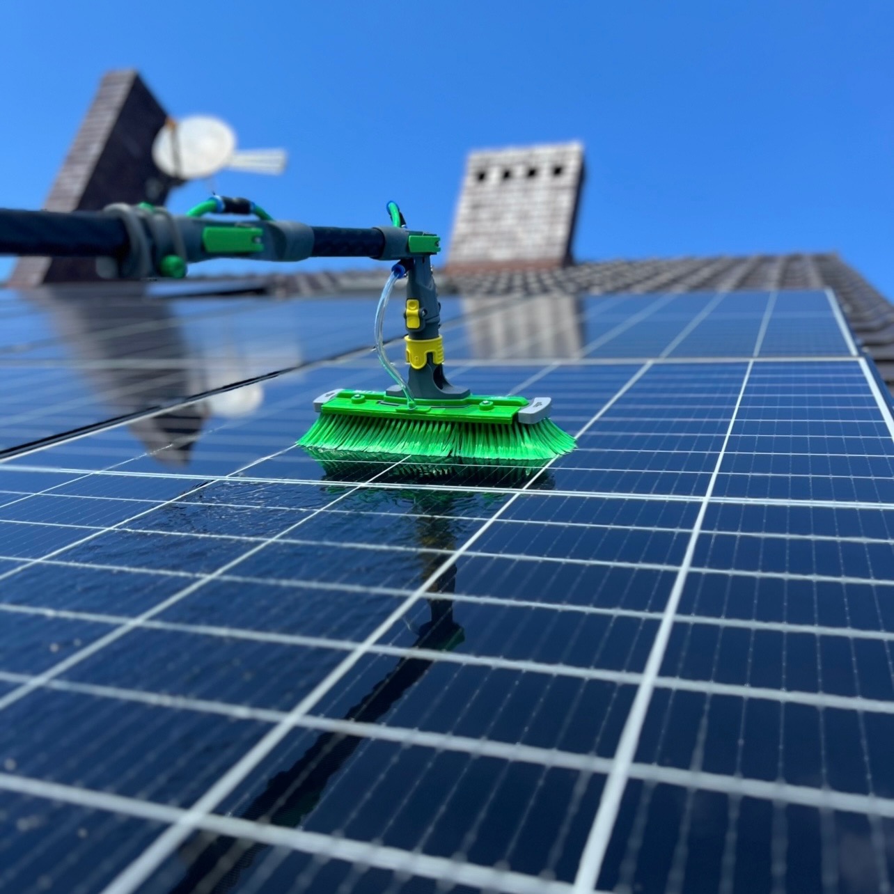 Szczotka teleskopowa z zielonym włosiem czyści panel słoneczny na dachu domu; w tle komin i błękitne niebo.