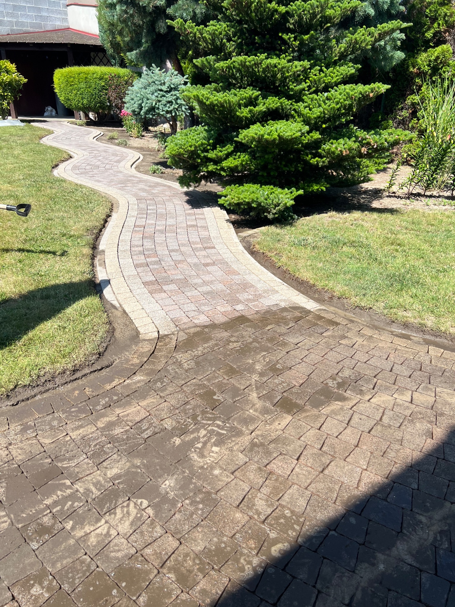 Mokry, kręty chodnik z kostki brukowej w ogrodzie, częściowo umyty, z widoczną różnicą w czystości. W tle zielona trawa i drzewa.