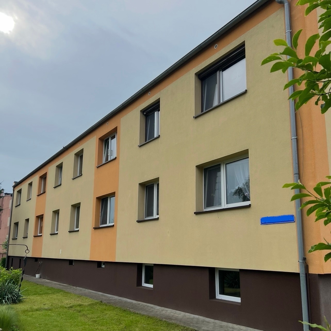 Odnowiona elewacja dwukondygnacyjnego budynku z oknami, pomalowana w odcieniach beżu i pomarańczy, z brązowym cokołem i zielonym trawnikiem przed budynkiem.