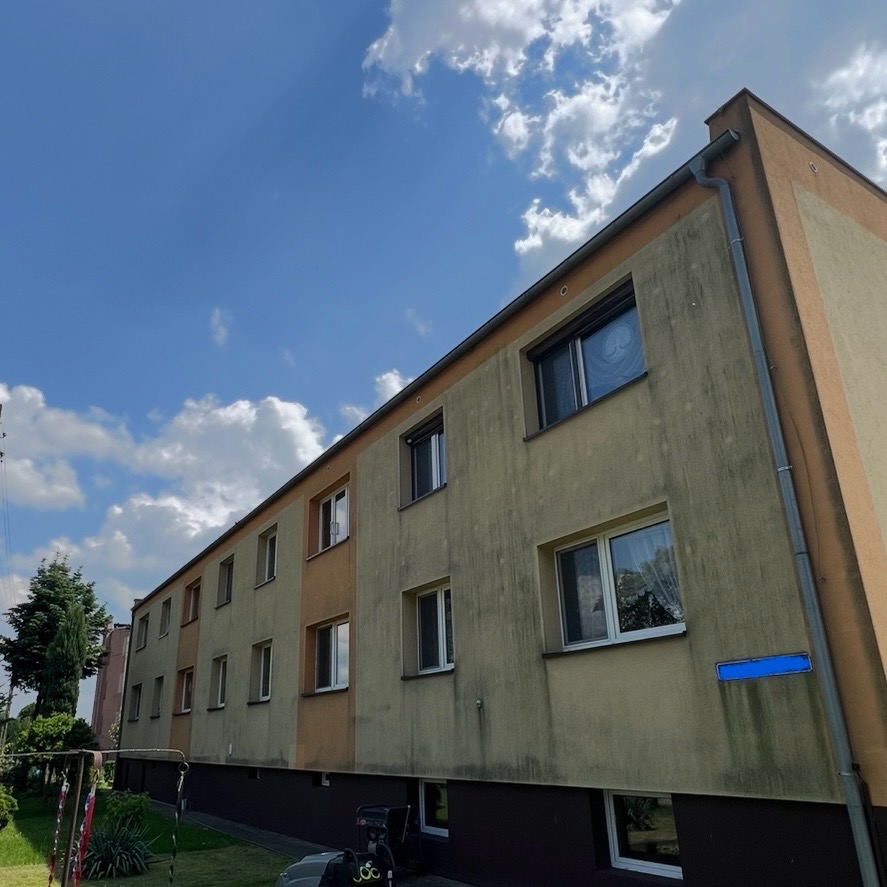 Wielorodzinny budynek z widocznymi zabrudzeniami na elewacji, sfotografowany pod kątem, z fragmentem niebieskiej tabliczki z nazwą ulicy. Słoneczny dzień.