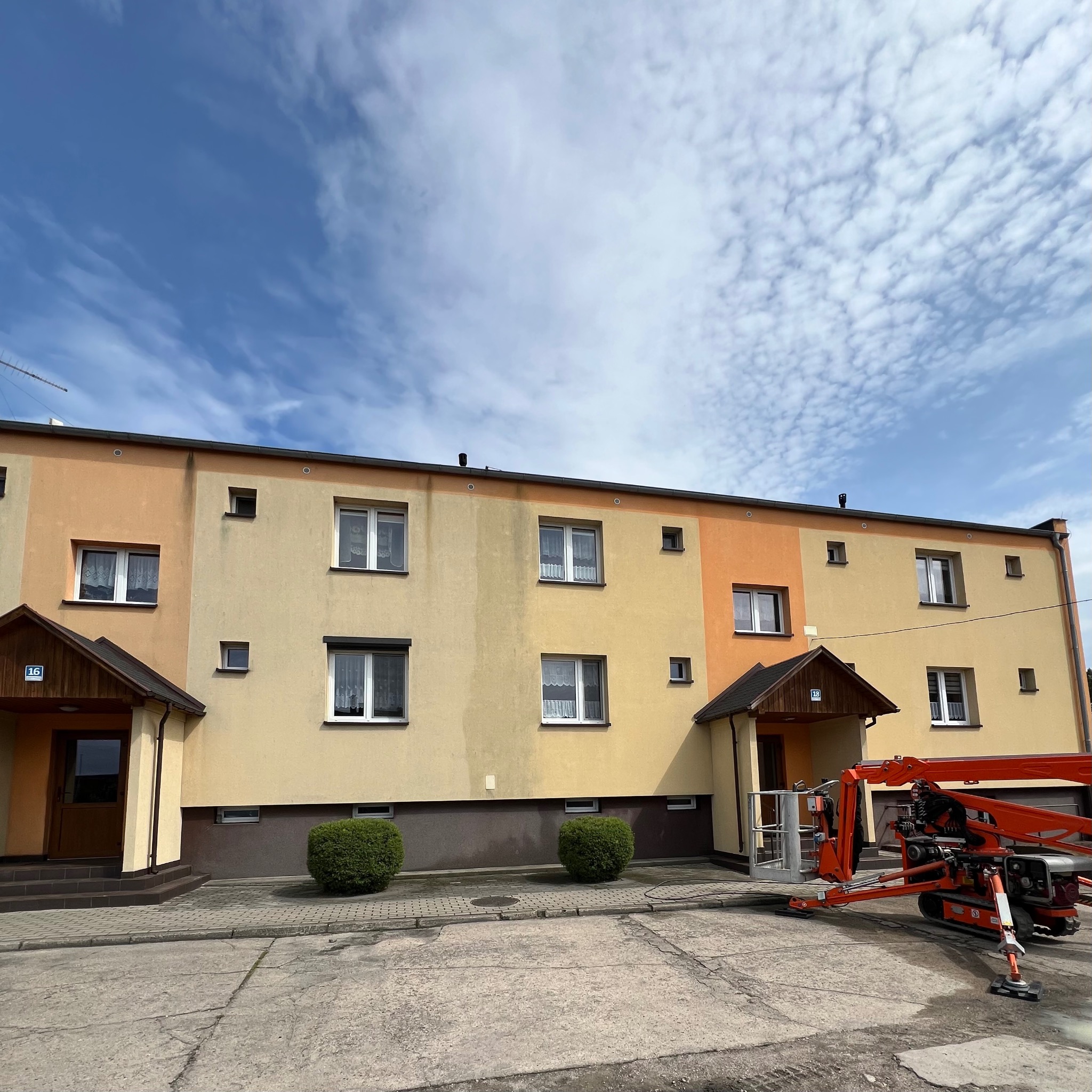 Wielorodzinny budynek z podnośnikiem koszowym, przygotowany do prac elewacyjnych. Widoczne zabrudzenia na fasadzie, sugerujące potrzebę czyszczenia lub malowania.