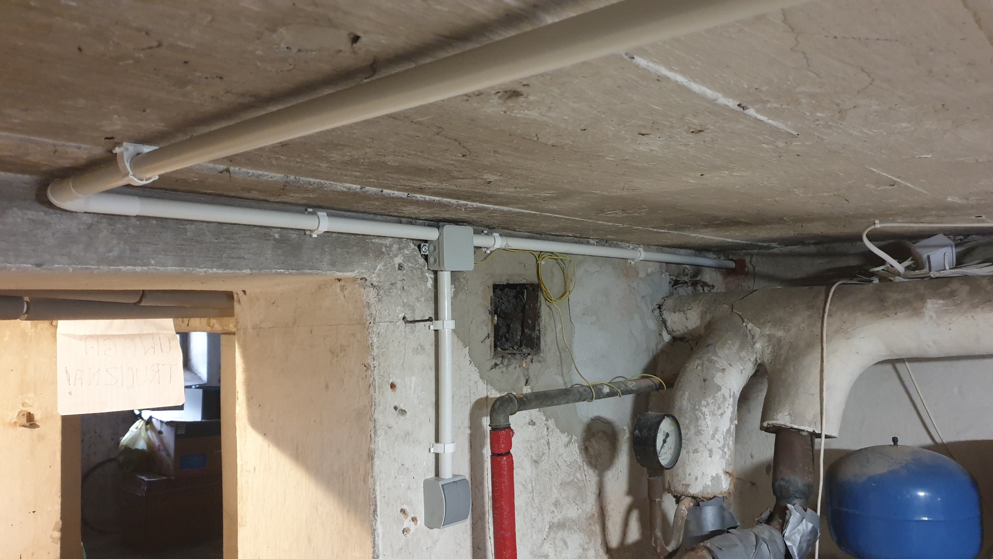 Instalacja natynkowa w piwnicy z rurami PCV, szarą puszką elektryczną i okablowaniem. Widoczne rury CO i fragment pieca. Remont starego budynku.