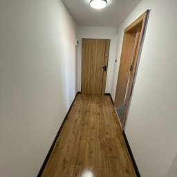FINITO WNĘTRZA Patryk Skoczylas - Korytarz po remoncie: drewnopodobne panele podłogowe, białe ściany i ościeżnice drzwiowe z drewnianym wykończeniem. Widok na dwie drewniane drzwi.