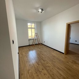 FINITO WNĘTRZA Patryk Skoczylas - Jasne wnętrze po remoncie: drewnopodobne panele, białe ściany. Drabina oparta o okno, widoczne listwy przypodłogowe i oświetlenie sufitowe. Puste pomieszczenie.