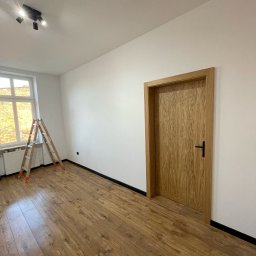 FINITO WNĘTRZA Patryk Skoczylas - Jasne panele podłogowe w pokoju po remoncie. Ściany pomalowane na biało, drewniane drzwi z czarną klamką. W rogu stoi drabina malarska.