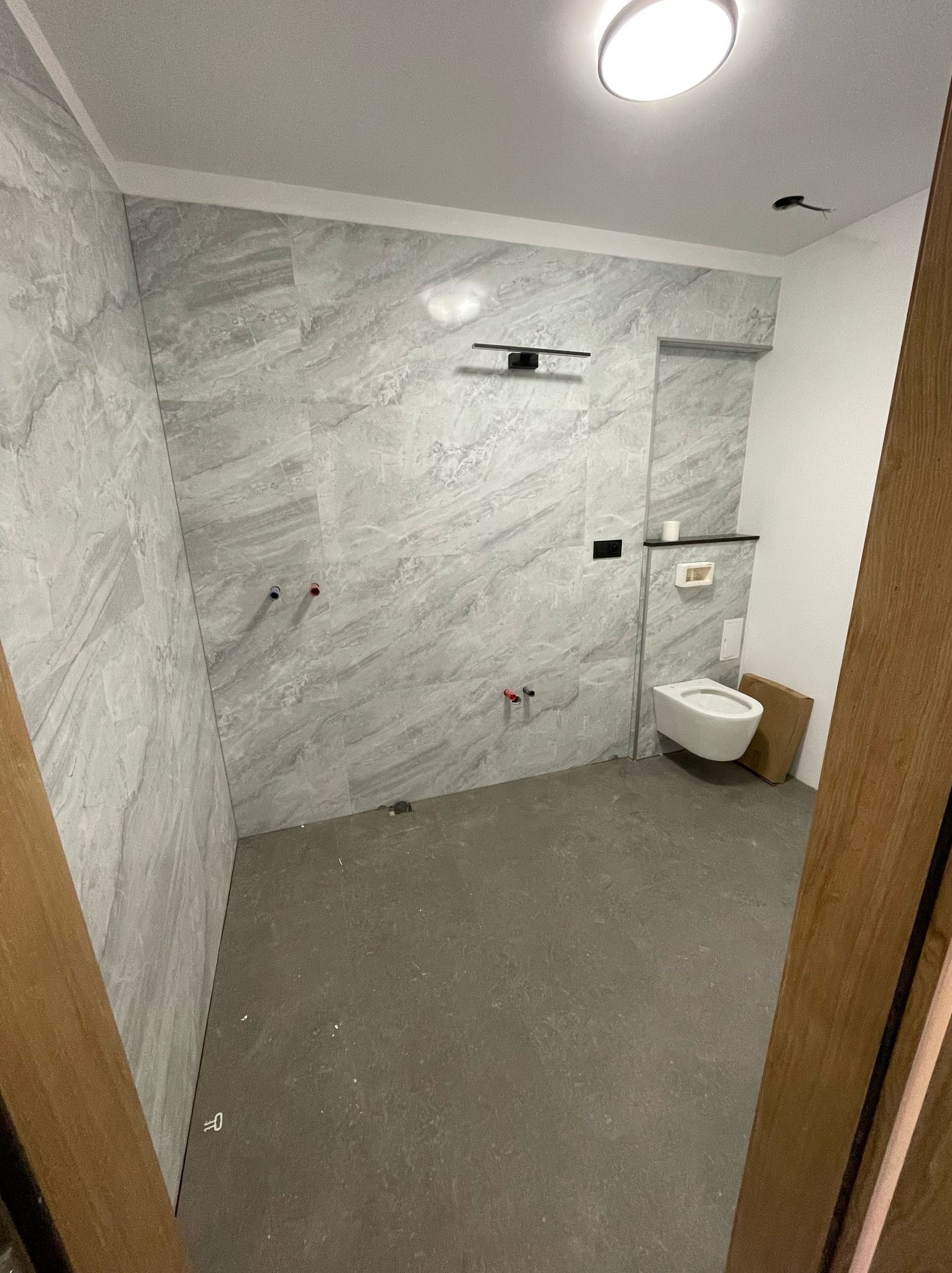 Wnętrze łazienki w trakcie wykończenia: szare płytki imitujące marmur na ścianach, częściowo zamontowana toaleta, surowa podłoga. Widoczne instalacje.