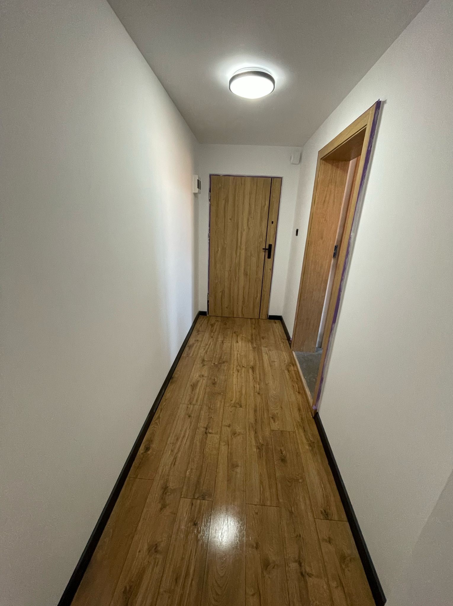 Korytarz po remoncie: drewnopodobne panele podłogowe, białe ściany i ościeżnice drzwiowe z drewnianym wykończeniem. Widok na dwie drewniane drzwi.