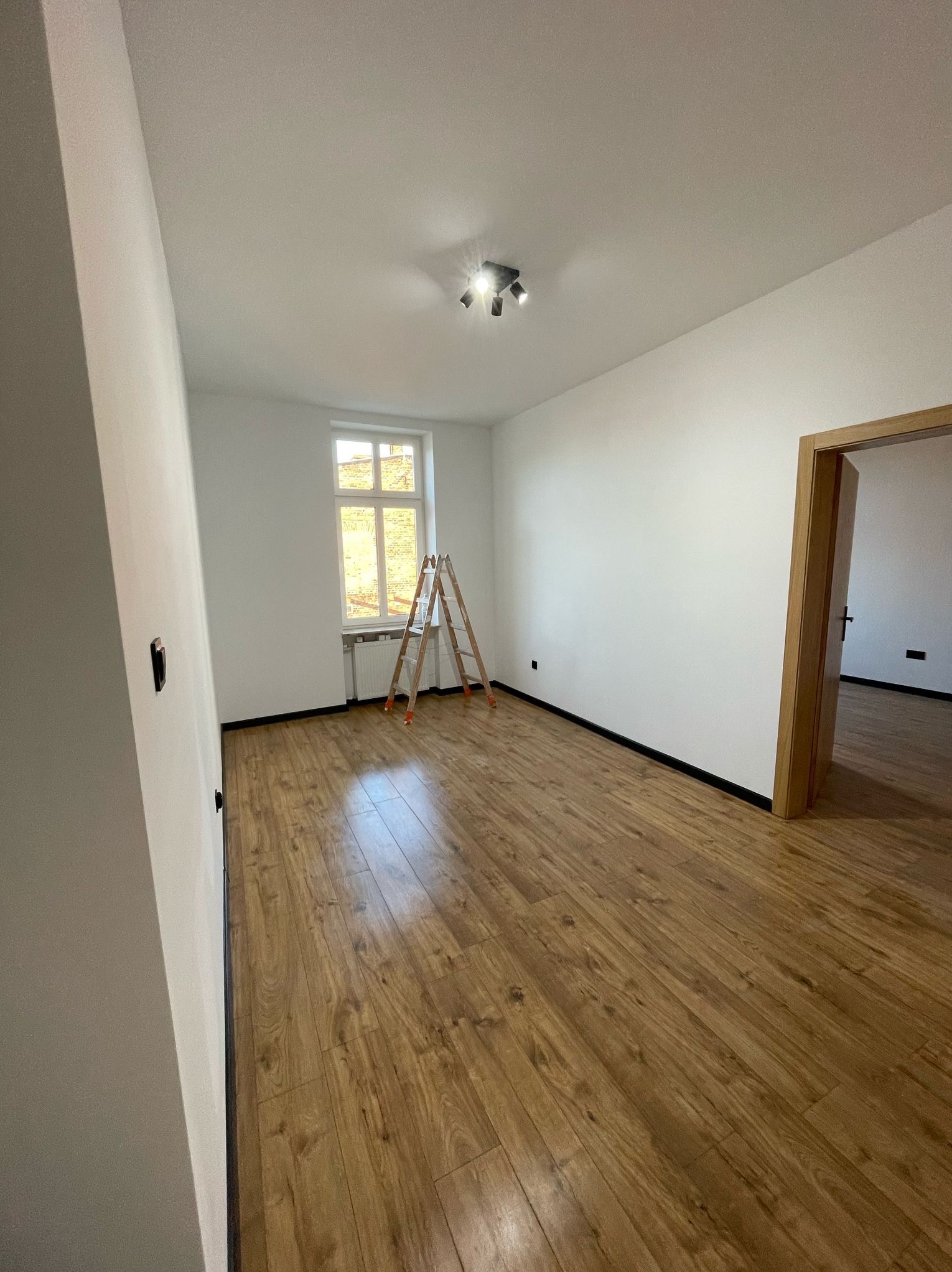 Jasne wnętrze po remoncie: drewnopodobne panele, białe ściany. Drabina oparta o okno, widoczne listwy przypodłogowe i oświetlenie sufitowe. Puste pomieszczenie.