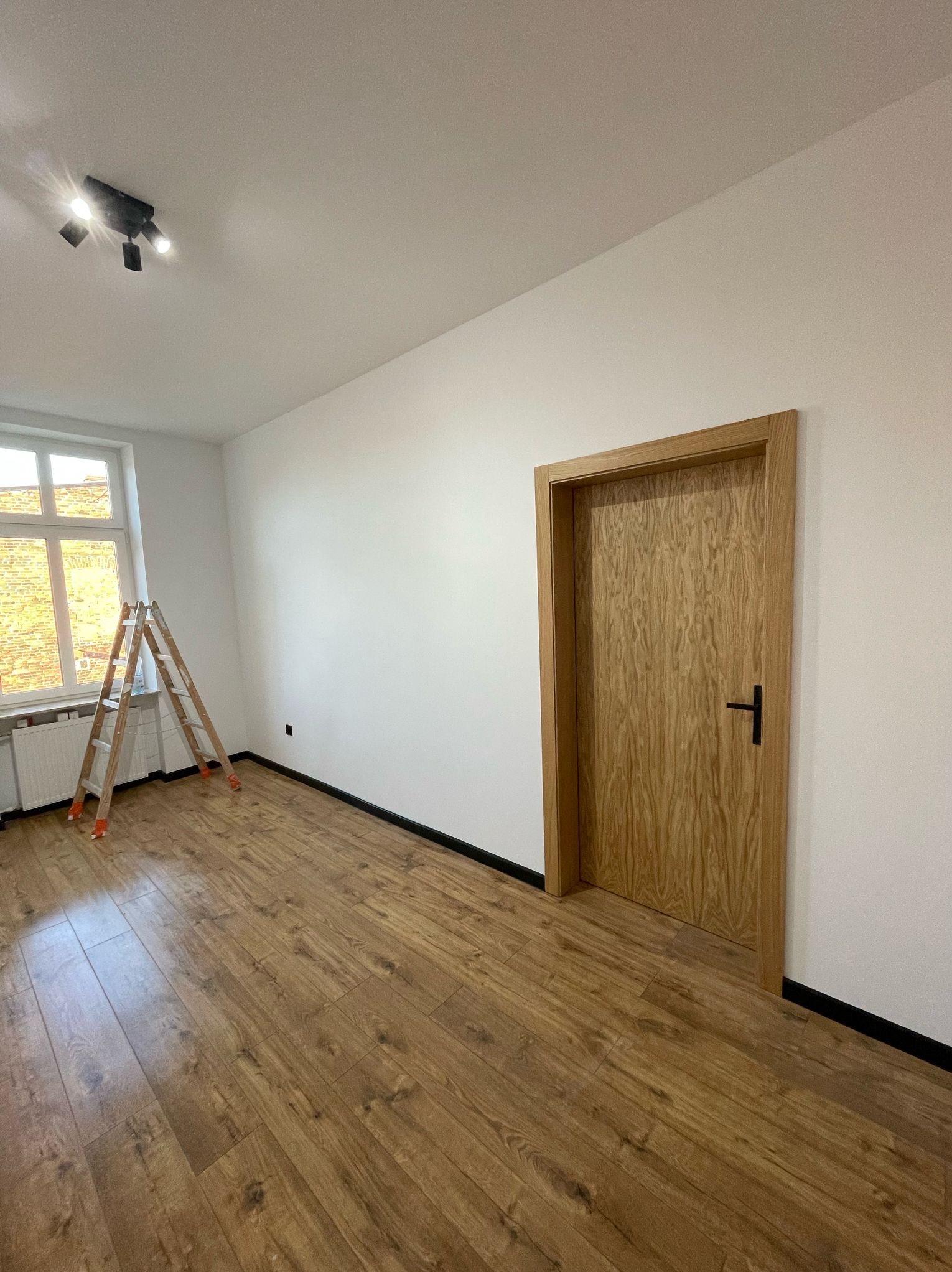 Jasne panele podłogowe w pokoju po remoncie. Ściany pomalowane na biało, drewniane drzwi z czarną klamką. W rogu stoi drabina malarska.