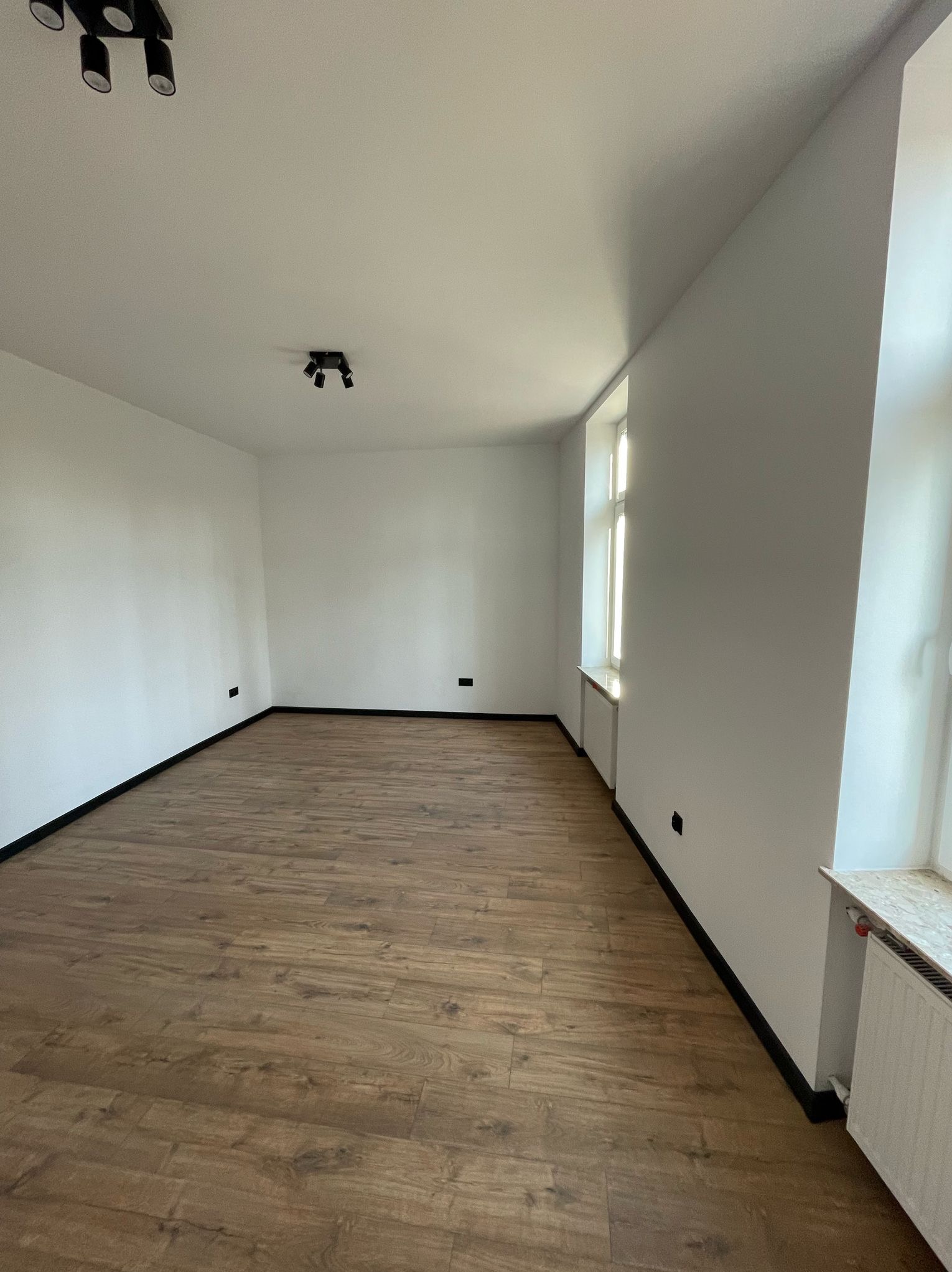 Jasne wnętrze po remoncie: brązowe panele, białe ściany z czarnymi listwami, okno z parapetem i grzejnik. Widoczne oświetlenie sufitowe.