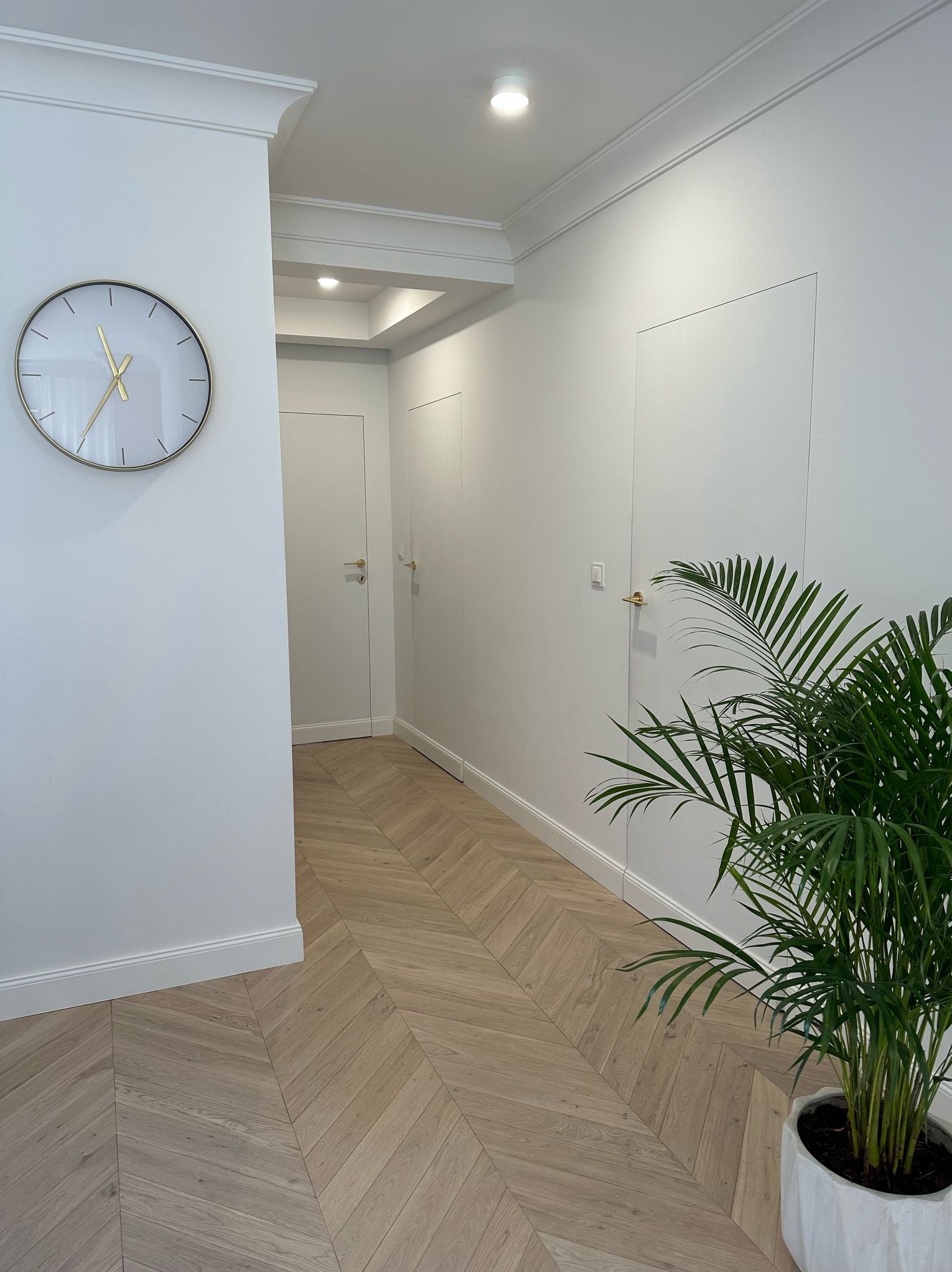 Biały korytarz z parkietem w jodełkę, ozdobiony zegarem i rośliną doniczkową. Minimalistyczne drzwi bez klamek wtapiają się w ścianę.