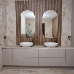 Projektowanie Wnętrz Nikola Kizyma - Nowoczesna łazienka z podwójną umywalką nablatową, owalnymi lustrami i drewnianą ścianą. Beżowe płytki podłogowe i subtelne oświetlenie.