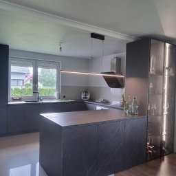 Projektowanie Wnętrz Nikola Kizyma - Nowoczesna kuchnia w odcieniach szarości z wyspą o marmurowym blacie i oświetleniem LED. Widoczna przeszklona witryna z kieliszkami i butelkami wina.