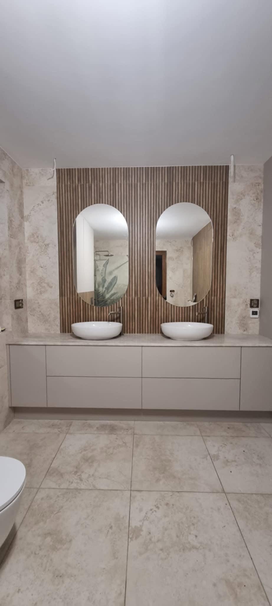 Nowoczesna łazienka z podwójną umywalką nablatową, owalnymi lustrami i drewnianą ścianą. Beżowe płytki podłogowe i subtelne oświetlenie.