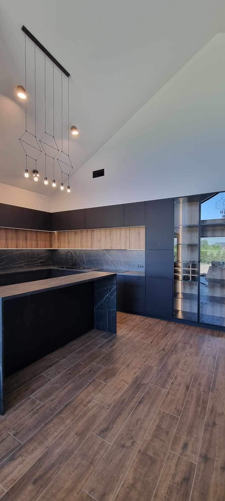 Nowoczesna kuchnia z wyspą, ciemnymi szafkami i blatem z marmuru, oświetlona wiszącymi lampami. Podłoga z płytek imitujących drewno. Duże okno z widokiem na zieleń.