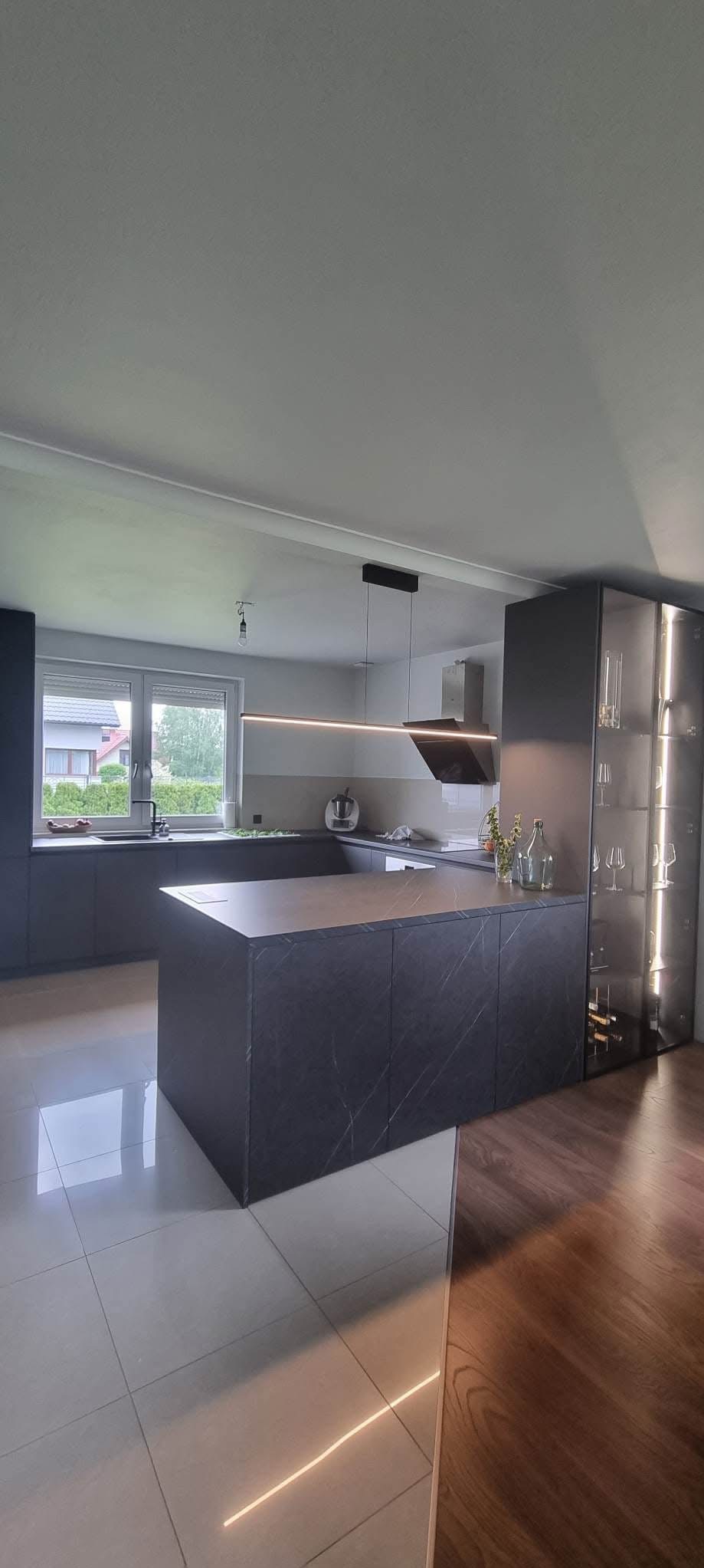 Nowoczesna kuchnia w odcieniach szarości z wyspą o marmurowym blacie i oświetleniem LED. Widoczna przeszklona witryna z kieliszkami i butelkami wina.