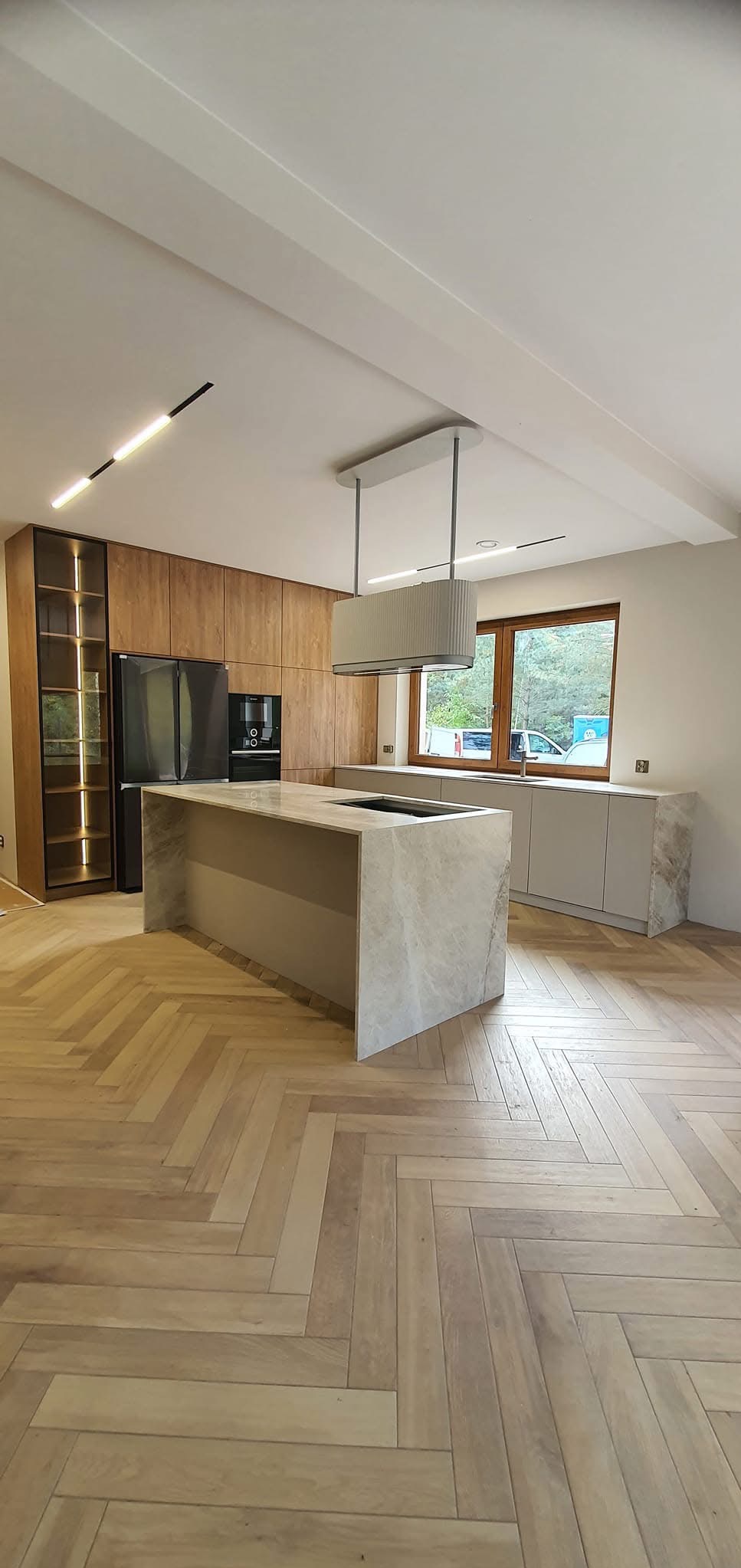 Nowoczesna kuchnia z wyspą wykończoną marmurem, parkiet w jodełkę, zabudowa drewniana z podświetlaną witryną i oknem. Minimalistyczny design i neutralna kolorystyka.