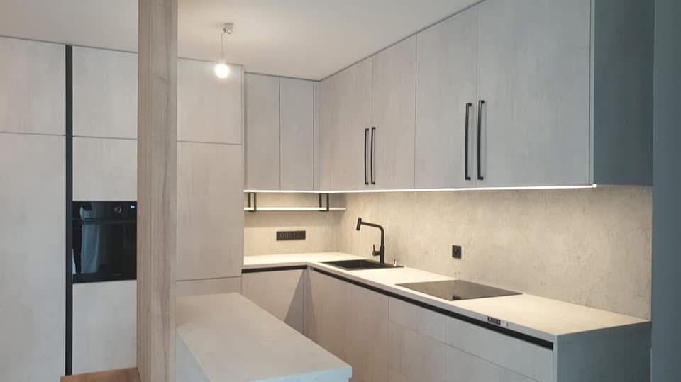 Nowoczesna kuchnia z szarymi szafkami, czarnymi uchwytami i oświetleniem LED pod szafkami. Widoczny piekarnik i czarny zlew. Minimalistyczny design.