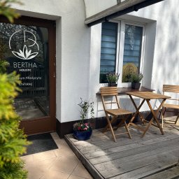 Bertha Holistic Centrum medycyny naturalnej - Wejście do centrum medycyny chińskiej Bertha Holistic z drewnianym stolikiem i krzesłami na zewnątrz, doniczkami z kwiatami i logo na szklanych drzwiach.