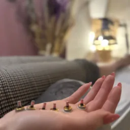 Dłoń z długimi paznokciami prezentuje sześć małych, okrągłych plastrów z wystającymi elementami, prawdopodobnie do terapii, w tle łóżko zabiegowe i dekoracyjne kwiaty.