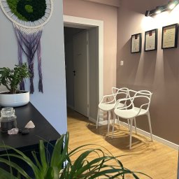 Bertha Holistic Centrum medycyny naturalnej - Wnętrze gabinetu z elementami dekoracyjnymi: makrama z symbolem Yin Yang z mchu, dwie białe krzesła, certyfikaty w ramkach na ścianie, rośliny doniczkowe i kryształy na komodzie.