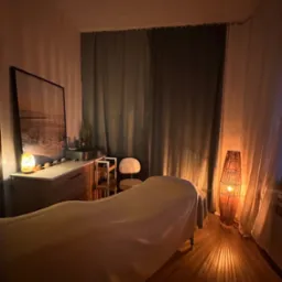 Przytulne wnętrze gabinetu masażu we Wrocławiu z łóżkiem do masażu przykrytym białym prześcieradłem, oświetlonym ciepłym światłem lampy z wikliny, z widocznym obrazem na ścianie i szafką z solną...