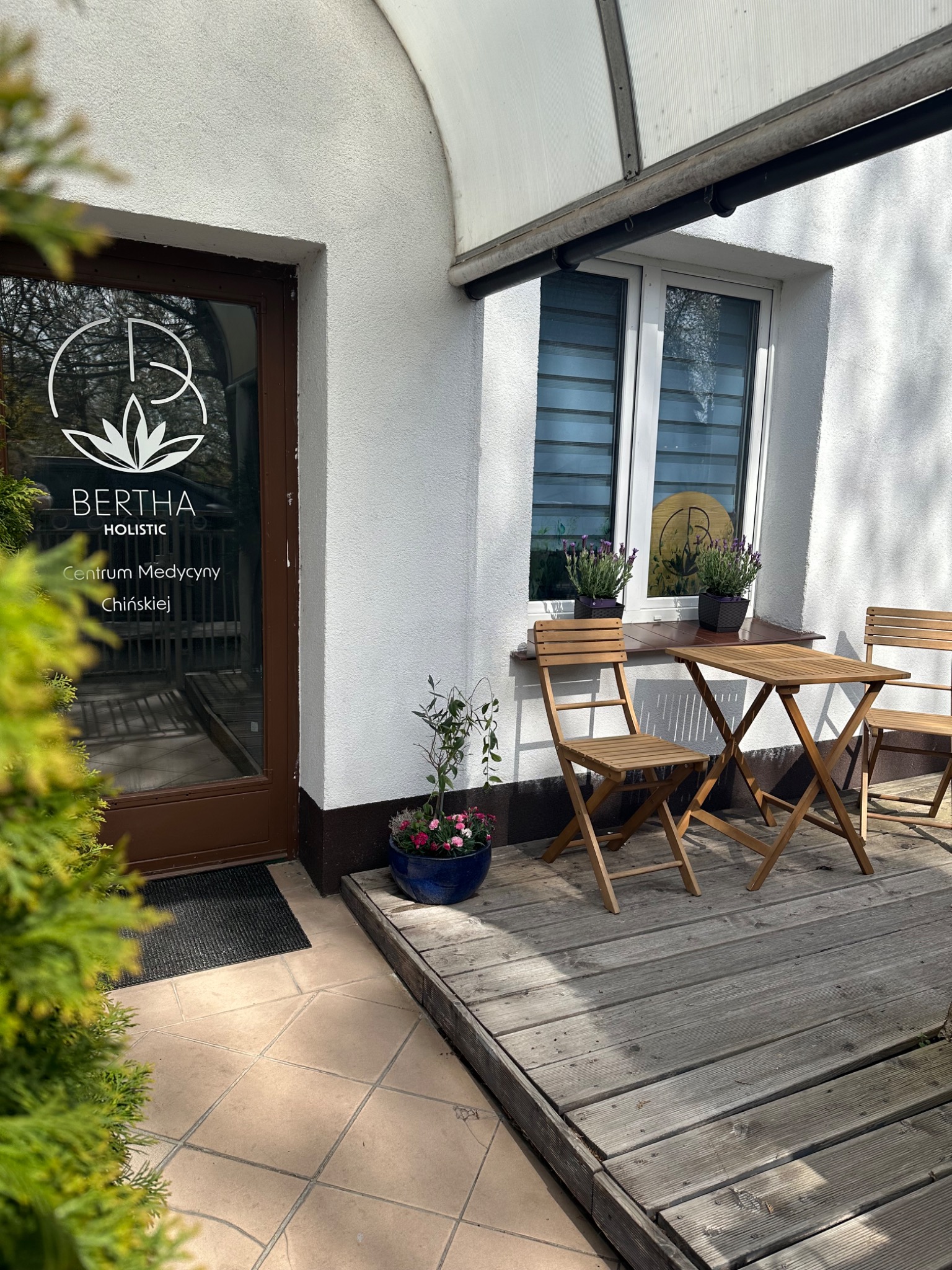 Wejście do centrum medycyny chińskiej Bertha Holistic z drewnianym stolikiem i krzesłami na zewnątrz, doniczkami z kwiatami i logo na szklanych drzwiach.
