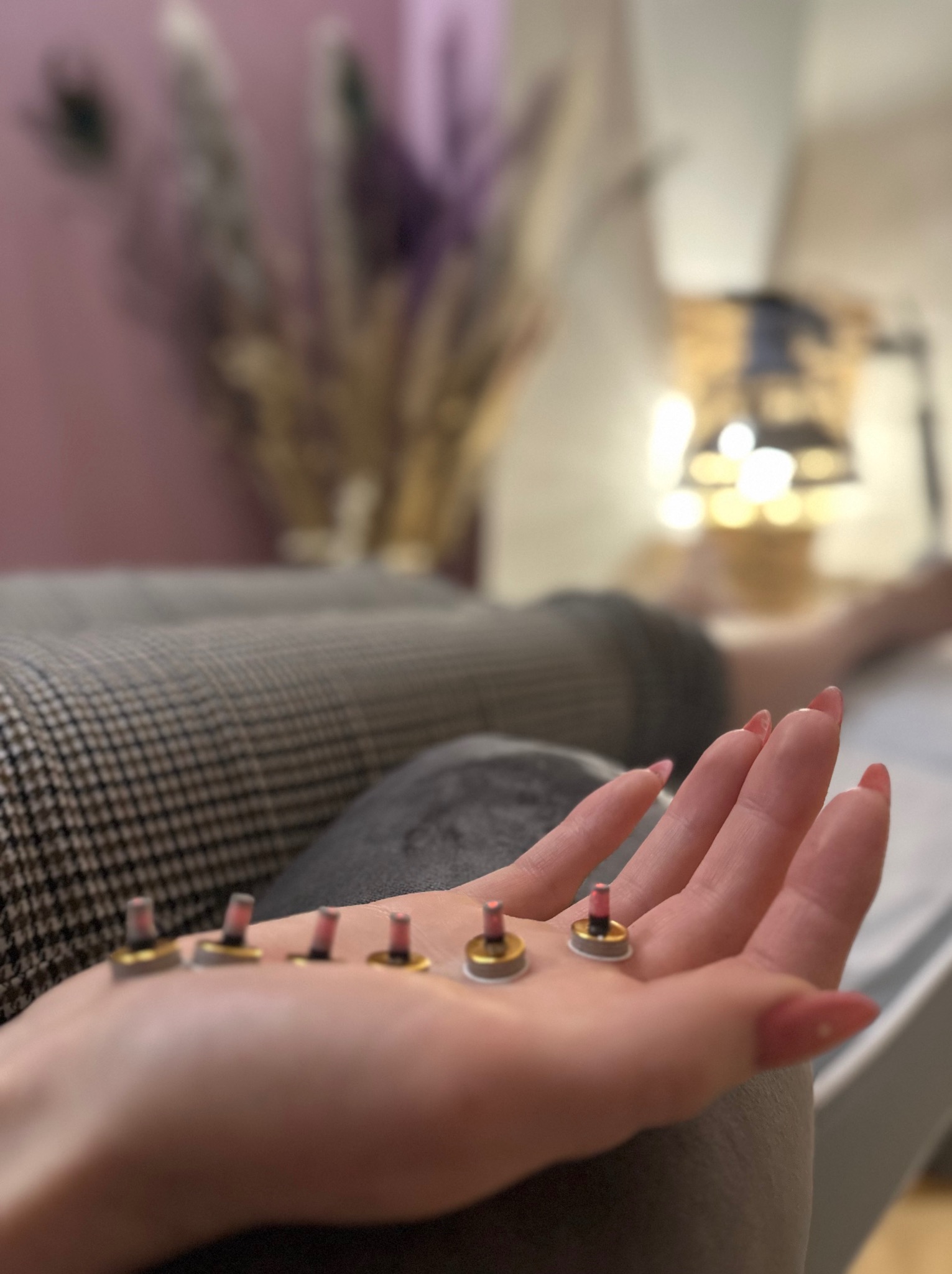 Dłoń z długimi paznokciami prezentuje sześć małych, okrągłych plastrów z wystającymi elementami, prawdopodobnie do terapii, w tle łóżko zabiegowe i dekoracyjne kwiaty.