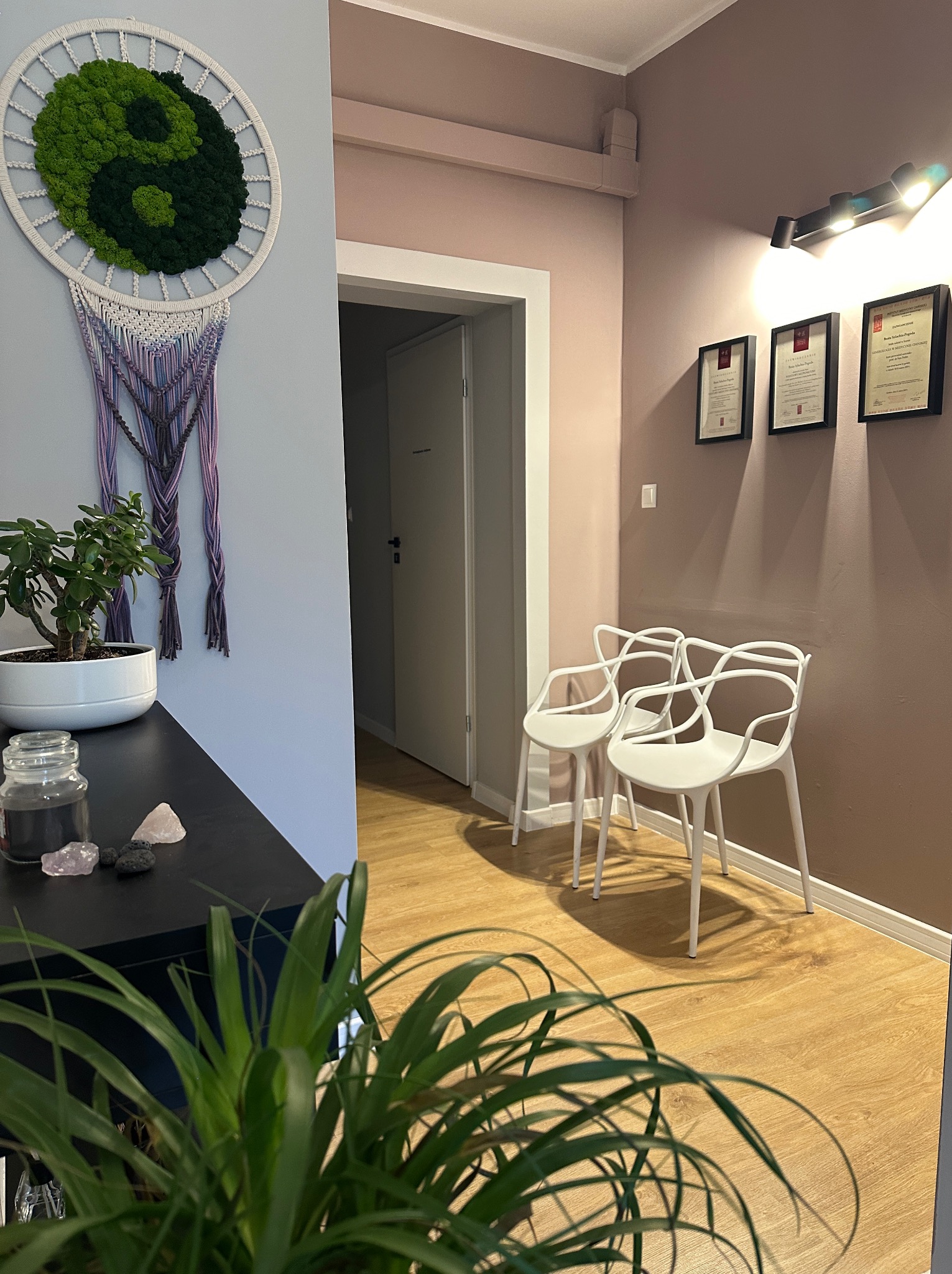 Wnętrze gabinetu z elementami dekoracyjnymi: makrama z symbolem Yin Yang z mchu, dwie białe krzesła, certyfikaty w ramkach na ścianie, rośliny doniczkowe i kryształy na komodzie.