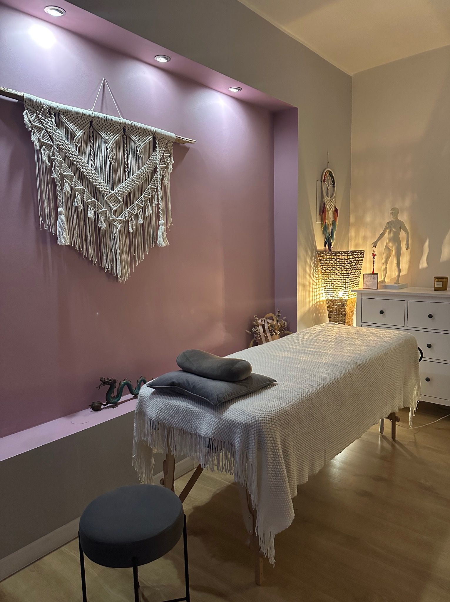 Pokój zabiegowy z łóżkiem przykrytym białym kocem, szare poduszki, dekoracja makrama na fioletowej ścianie, figurka smoka i statuetka człowieka na komodzie.