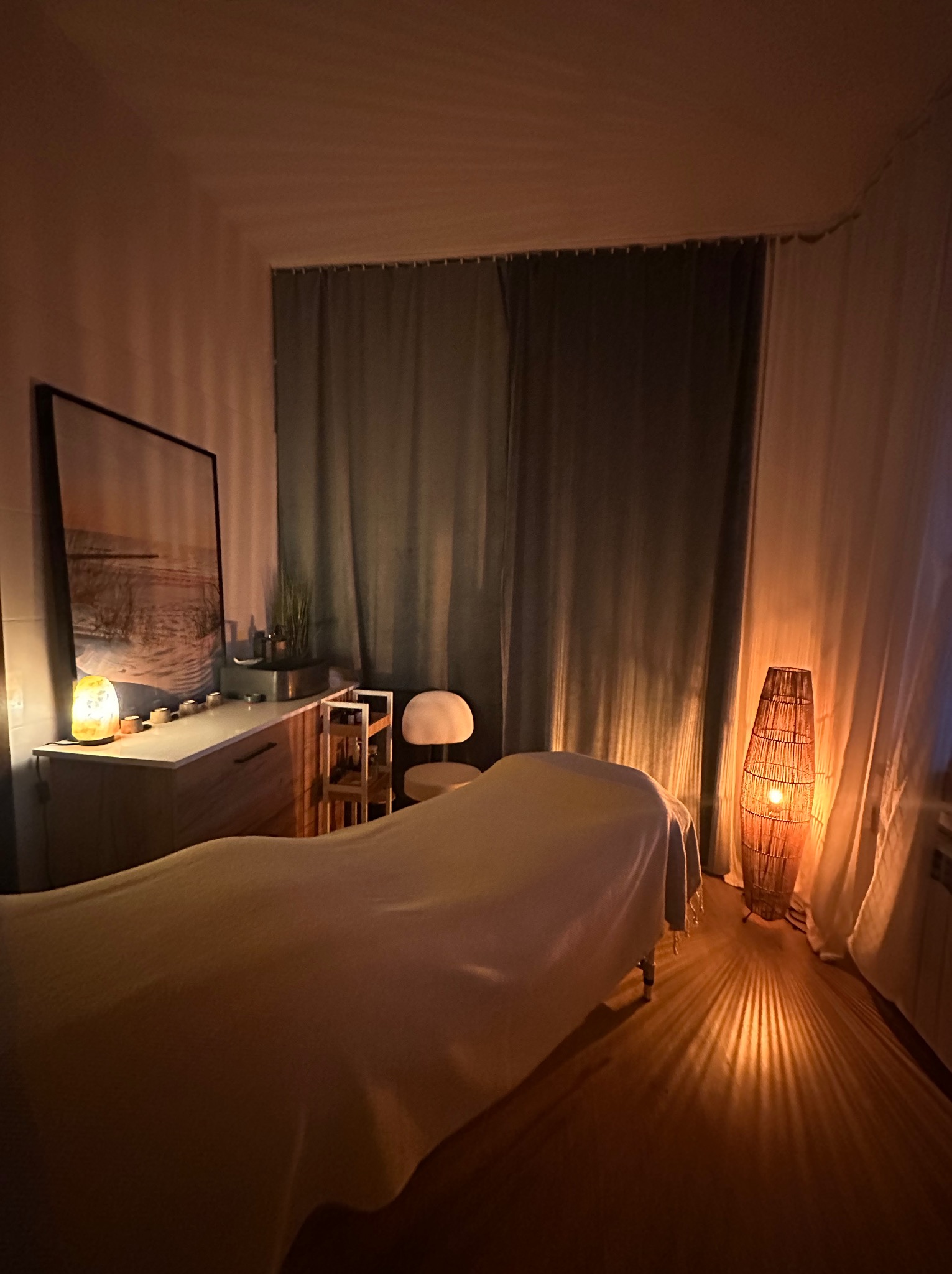 Przytulne wnętrze gabinetu masażu we Wrocławiu z łóżkiem do masażu przykrytym białym prześcieradłem, oświetlonym ciepłym światłem lampy z wikliny, z widocznym obrazem na ścianie i szafką z solną...