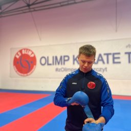 Bartosz Kędzierski - Młody mężczyzna zakłada niebieskie rękawice do karate na tle maty i baneru klubu sportowego KS Olimp.