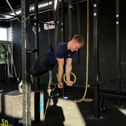 Bartosz Kędzierski - Mężczyzna w okularach wykonuje ćwiczenie na drewnianych kółkach gimnastycznych w siłowni, z obciążeniem w postaci kettlebell zawieszonego na łańcuchu.