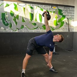 Bartosz Kędzierski - Mężczyzna w okularach wykonuje ćwiczenie z kettlebellem nad głową w siłowni z graffiti na ścianie.