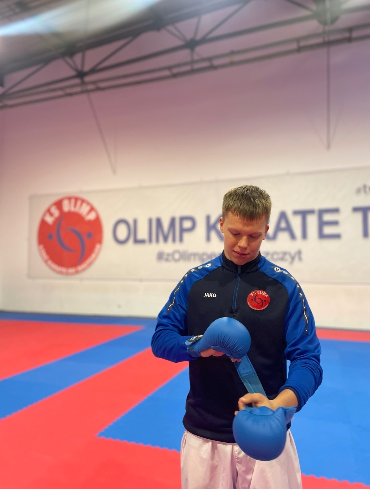 Młody mężczyzna zakłada niebieskie rękawice do karate na tle maty i baneru klubu sportowego KS Olimp.