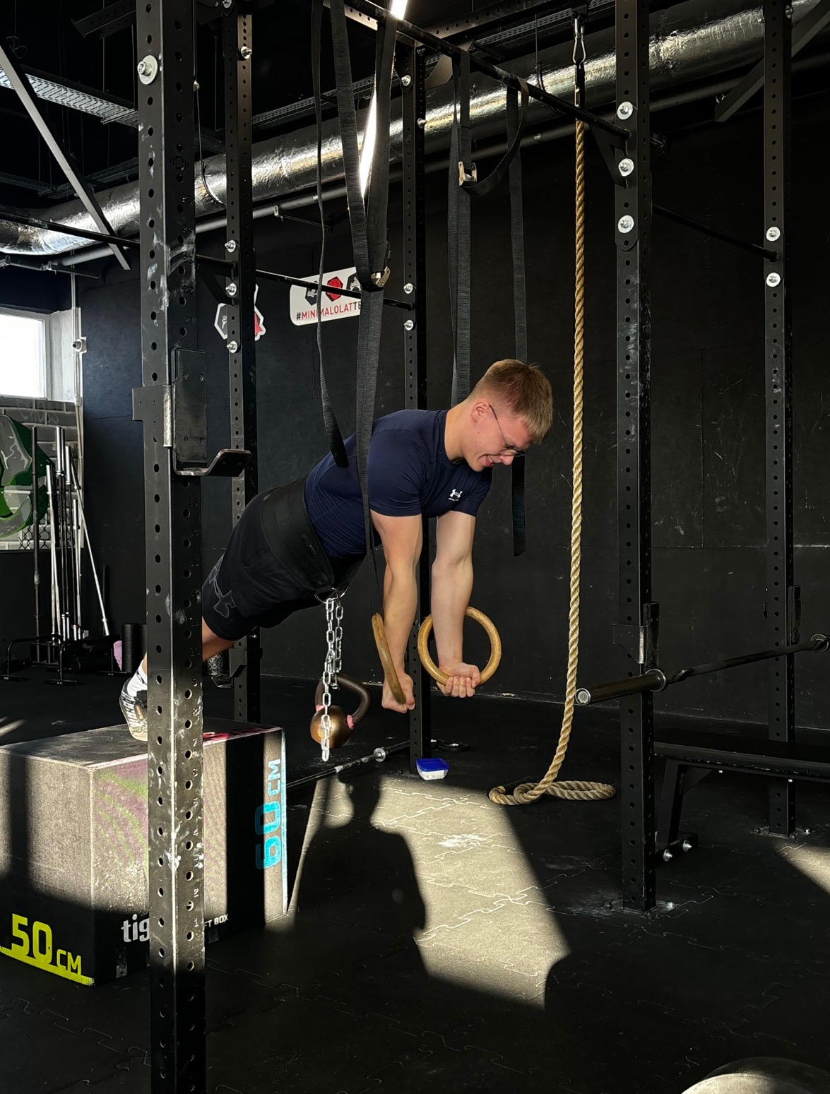 Mężczyzna w okularach wykonuje ćwiczenie na drewnianych kółkach gimnastycznych w siłowni, z obciążeniem w postaci kettlebell zawieszonego na łańcuchu.