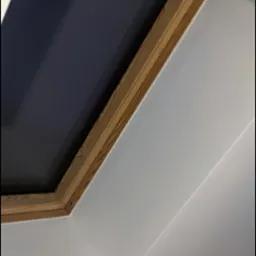 Okno dachowe z drewnianą ramą, widziane od wewnątrz. Białe ściany i sufit. Widoczny mechanizm rolety. Ujęcie z dołu, skierowane w górę.