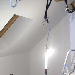 OsBud - Poddasze w trakcie prac wykończeniowych: widoczne kable, oświetlenie robocze i drabina. Ściany pomalowane na biało, skosy dachu. Surowy stan przed wykończeniem.